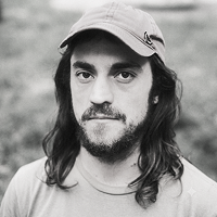 A black and white photo of a man with long dark hair, a beard, and wearing a ball cap.